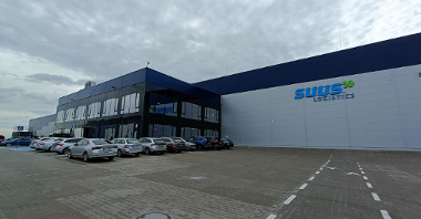 The photo shows the warehouse building of SUUS Logistics. There is a glass entrance in front of the warehouse. Passenger cars are parked in the car park.