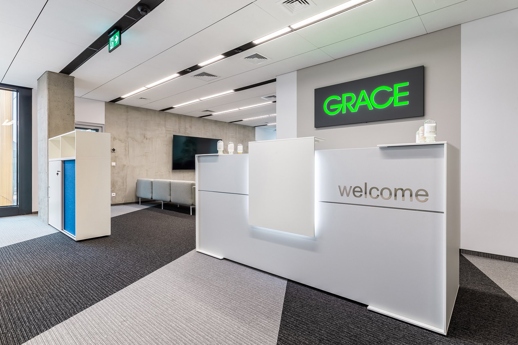 The photo shows a white wall, displaying the Grace company logo. In front of it is a white reception desk with the words 'Welcome'. - grafika artykułu