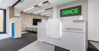 The photo shows a white wall, displaying the Grace company logo. In front of it is a white reception desk with the words 'Welcome'.