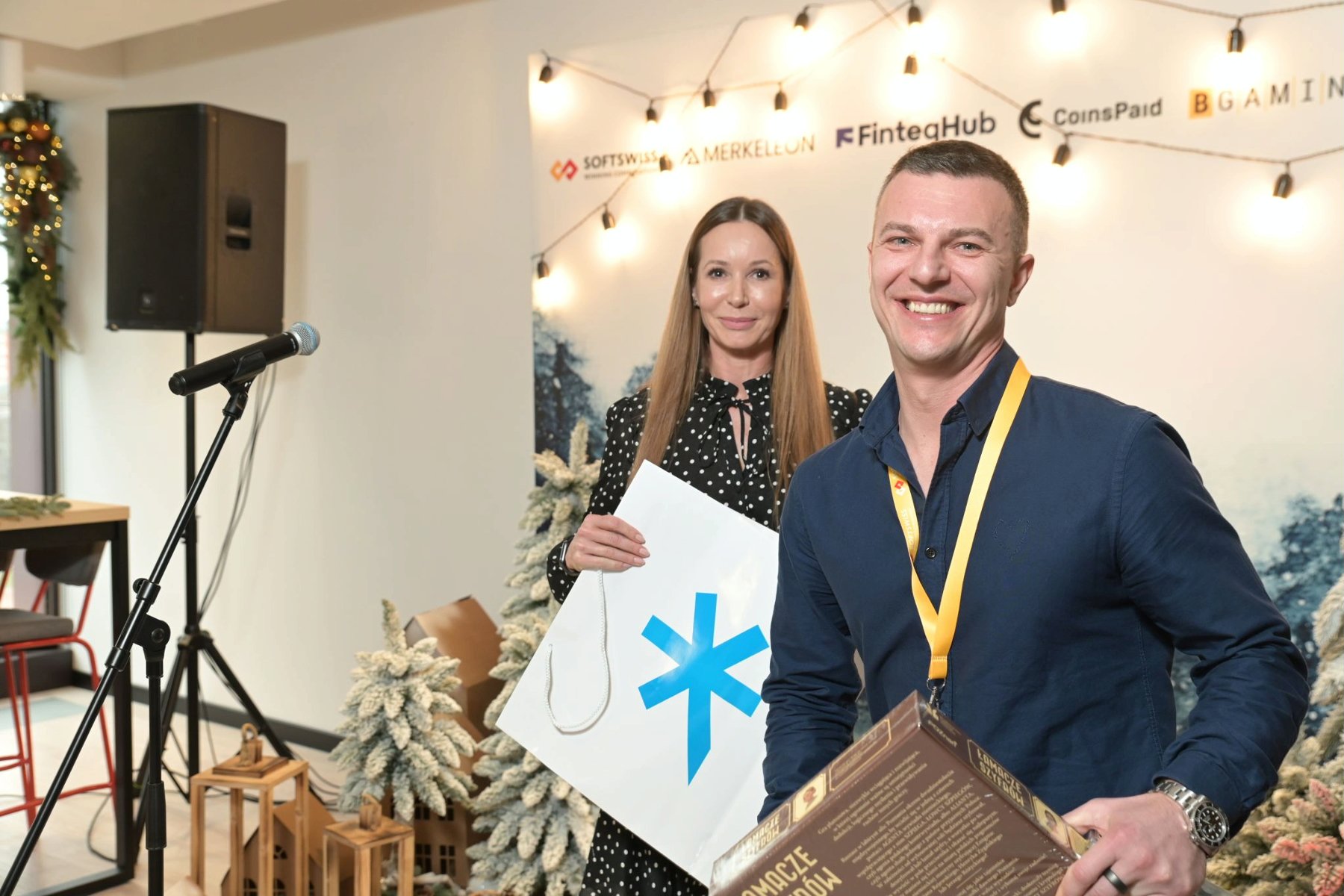 The photo shows Ivan Montik, founder of SOFTSWISS. The man is smiling, holding the board game Code Breakers. Behind him is the director of the Investor Relations Department, Katja Lozina. In the background is a festive wall with company logos. - grafika artykułu