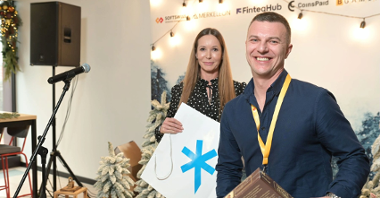 The photo shows Ivan Montik, founder of SOFTSWISS. The man is smiling, holding the board game Code Breakers. Behind him is the director of the Investor Relations Department, Katja Lozina. In the background is a festive wall with company logos.