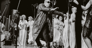 A black and white photo from a concert: a man in sunglasses and a black leather coat performing on stage, behind him women in long white dresses.