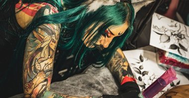 A tattooed girl with green hair and heavy makeup is tattooing a client.