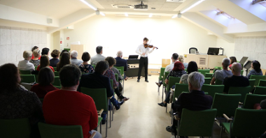 The photo shows a man playing the violin in from of people seating