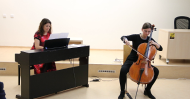 The photo shows a lady playing a bass and another lady playing a piano